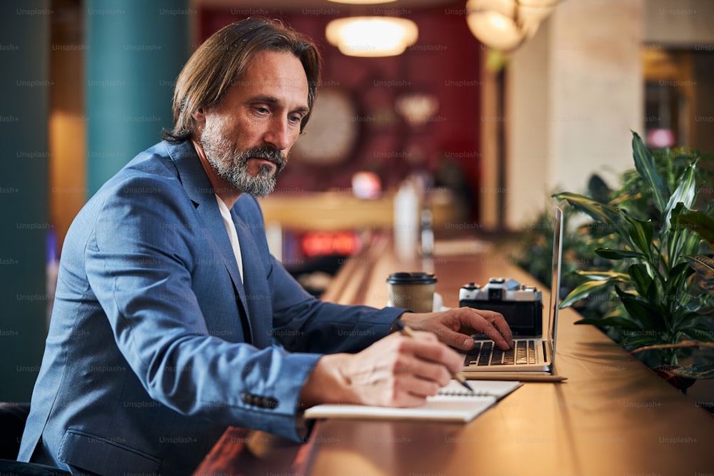 Konzentrierter reifer Herr, der sich Notizen macht, während er vor einem Laptop an einem bequemen Schreibtisch sitzt