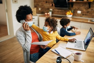 African American working mother talking on the phone while daughter is reminding her that she has online meeting over laptop at home. They are wearing protective face masks due to coronavirus pandemic.