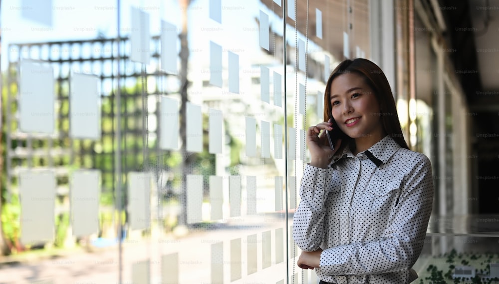 A cheerful businesswoman is talking on phone while standing near by a window in modern office.