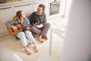 Beau jeune homme assis sur le sol et souriant pendant que sa charmante petite amie joue de la guitare