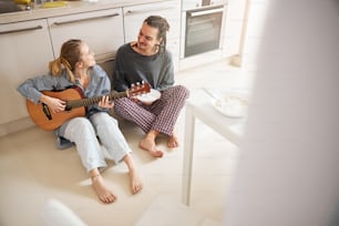 Handsome young man sitting on the floor and smiling while his charming girlfriend playing guitar