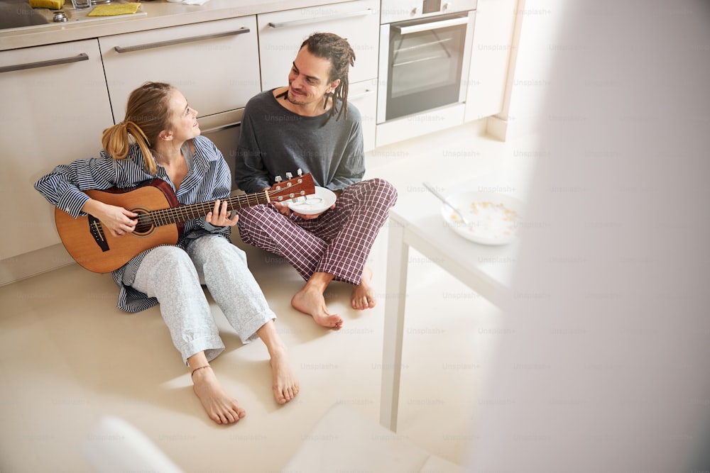 ハンサムな若い男は床に座って微笑みながら、彼の魅力的なガールフレンドがギターを弾いています