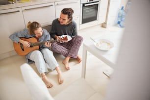 Beautiful female musician using acoustic musical instrument while sitting on the floor beside young man