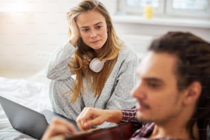 Charming lady in pajamas looking at boyfriend with love and smiling while man playing guitar