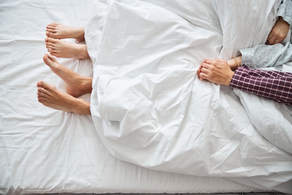Close up of barefoot boyfriend and girlfriend resting under white blanket in bedroom