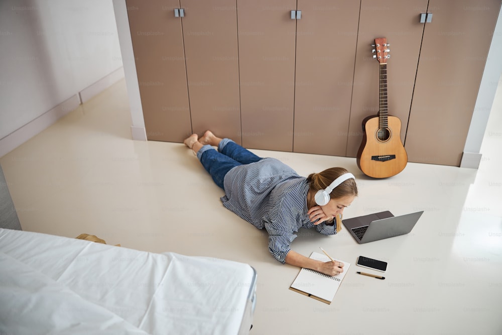 Charming young woman writing songs while lying on the floor with laptop and cellphone
