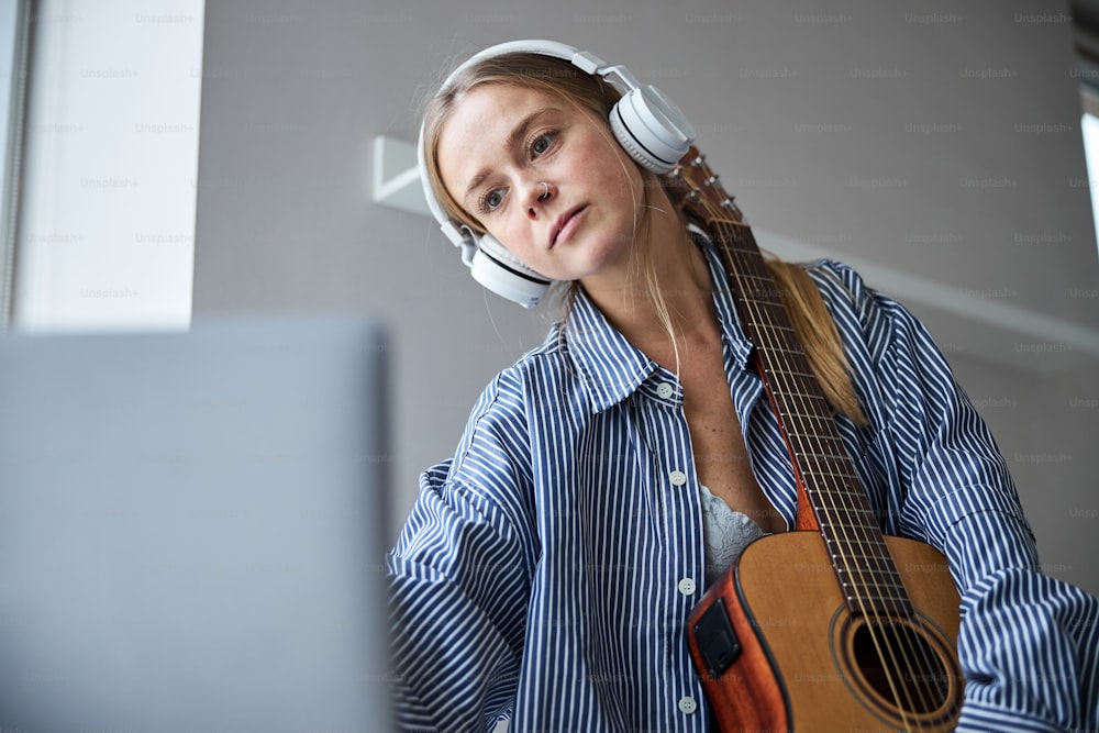 Beautiful female musician in headphones holding acoustic musical instrument and surfing the internet on notebook