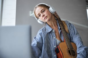 Beautiful female musician in headphones holding acoustic musical instrument and surfing the internet on notebook
