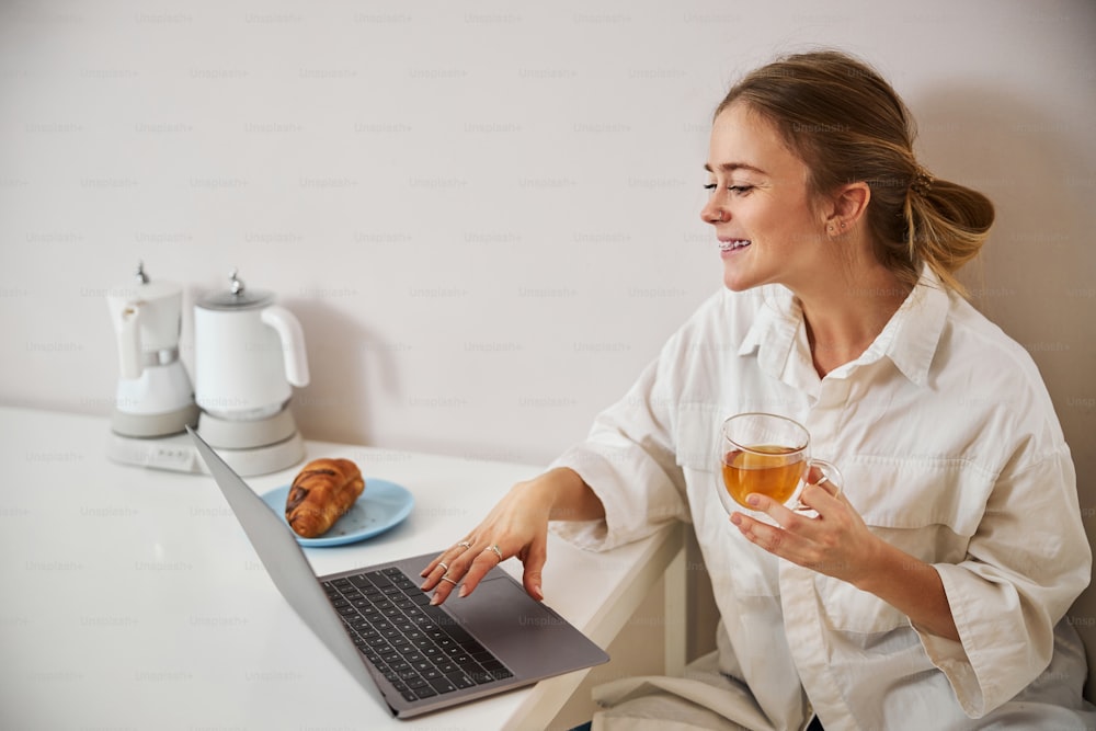 Joyful pretty lady holding cup of herbal tea and smiling while sitting at the table with modern laptop