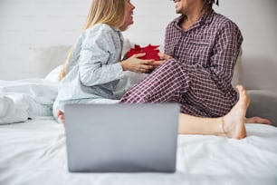 Encantadora joven en pijama mirando a su novio y sonriendo mientras acepta una caja de regalo roja