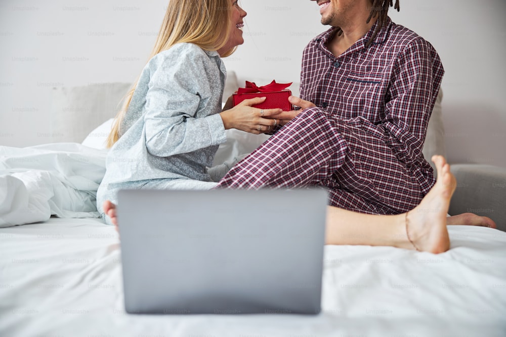 Charming young woman in pajamas looking at boyfriend and smiling while accepting red gift box