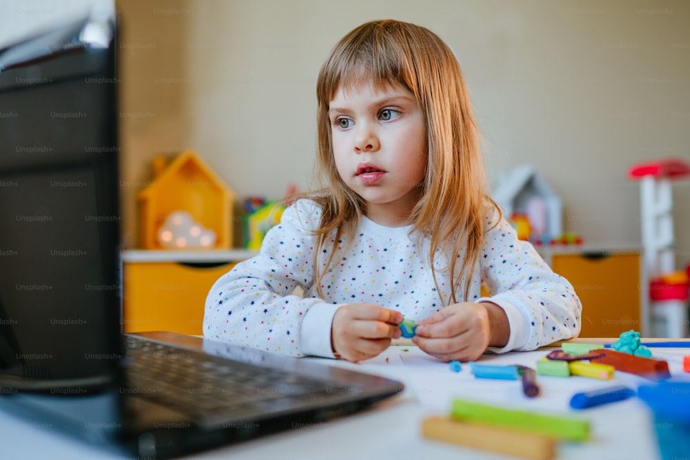 어린 소녀는 실내에서 노트북으로 온라인 학습 수업을 보고 다채로운 점토를 성형합니다. 원격 가정 학습 개념입니다.