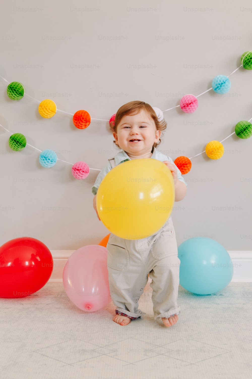 Funny Caucasian baby boy celebrating his first birthday. Excited child kid toddler playing with colorful balloons. Celebration of event or party indoors at home. Happy birthday lifestyle concept.