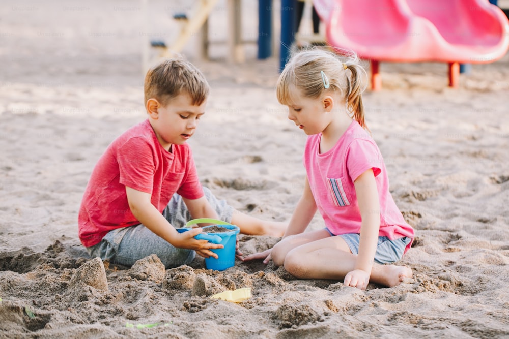 Due bambini caucasici seduti in sabbiera che giocano con giocattoli da spiaggia. Bambina e ragazzi che si divertono insieme nel parco giochi. Attività estiva all'aperto per bambini. Tempo libero, stile di vita, infanzia.