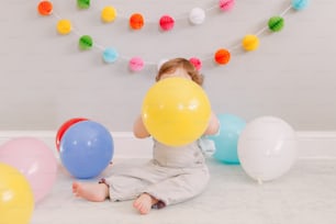 Funny Caucasian baby boy celebrating his first birthday. Child kid toddler sitting on floor with colorful balloons. Celebration of event or party indoors at home. Happy birthday lifestyle concept.