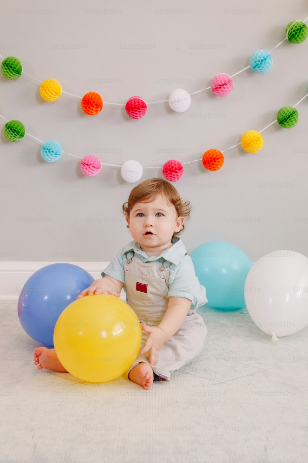 Funny Caucasian baby boy celebrating his first birthday. Child kid toddler sitting on floor with colorful balloons. Celebration of event or party indoors at home. Happy birthday lifestyle concept.