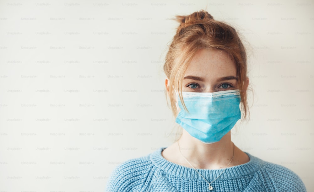 Red haired caucasian woman with freckles is wearing a medical mask and looking at camera on a white studio wall with free space