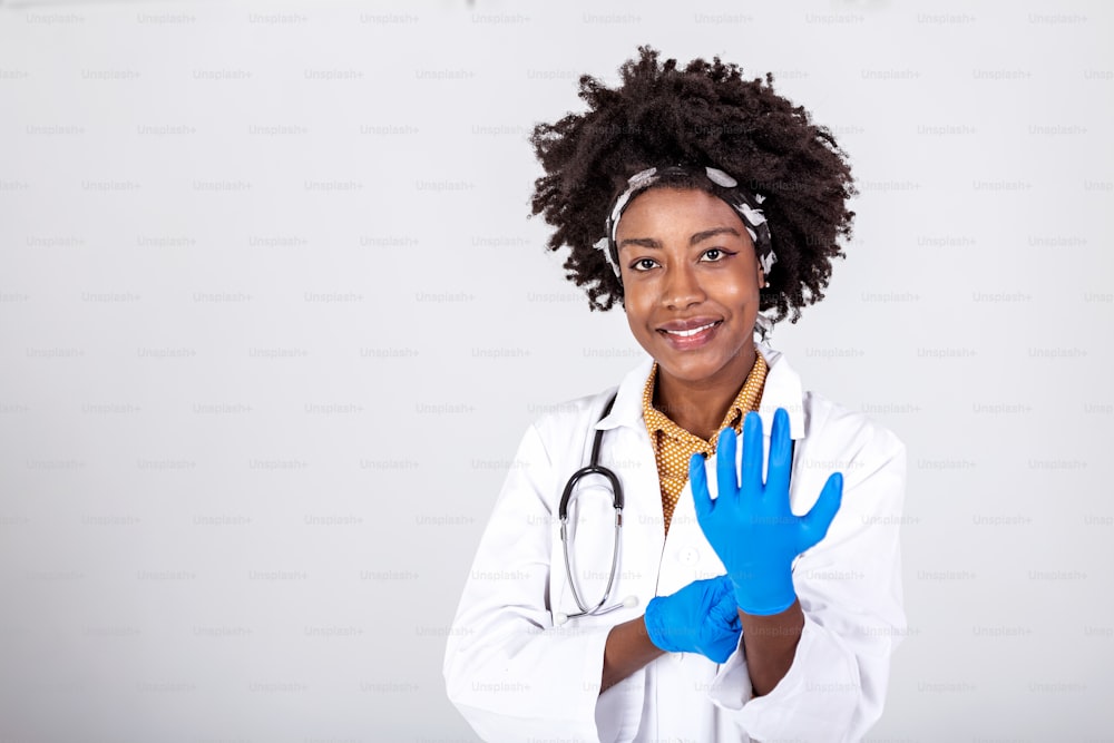 Retrato de una atractiva joven doctora con bata blanca poniéndose guantes médicos. Retrato del médico sonriente alegre en uniforme blanco de pie con las manos cruzadas sobre fondo gris