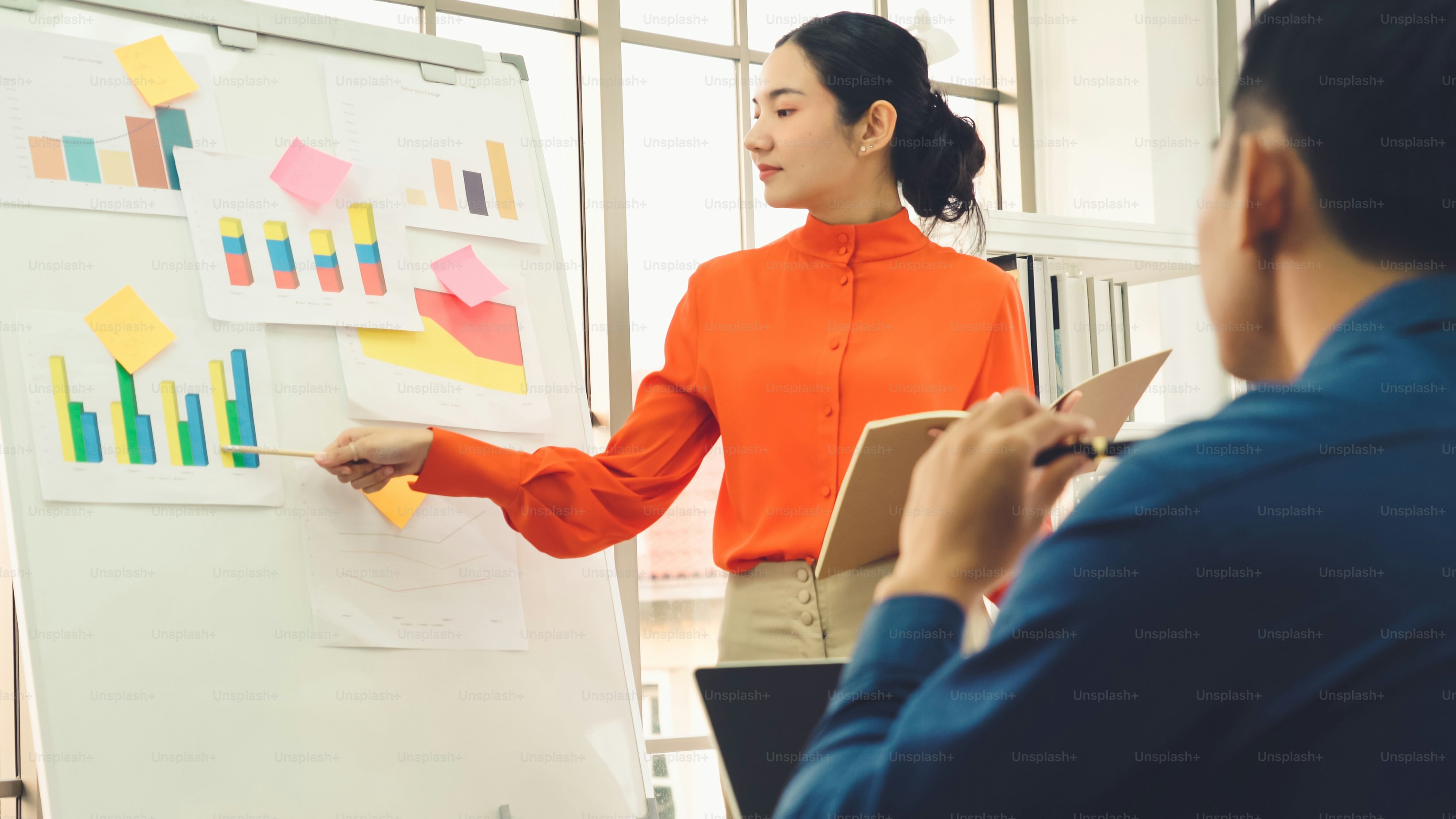 Young woman explains business data on white board in casual office room . The confident Asian businesswoman reports information progress of a business project to partner to determine market strategy .