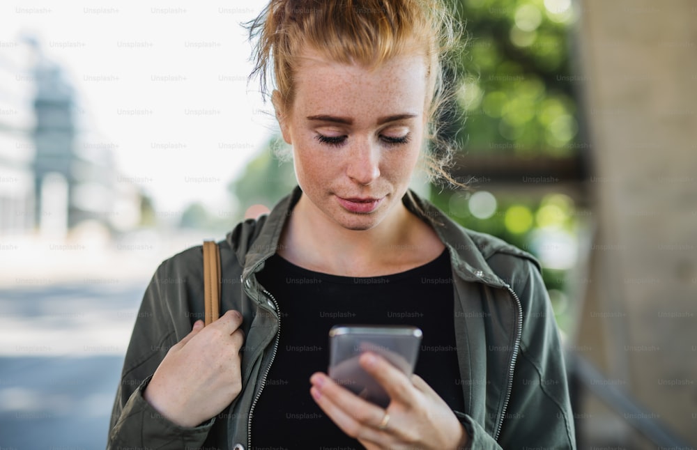 Front view portrait of young woman with red hair outdoors in town, using smartphone.