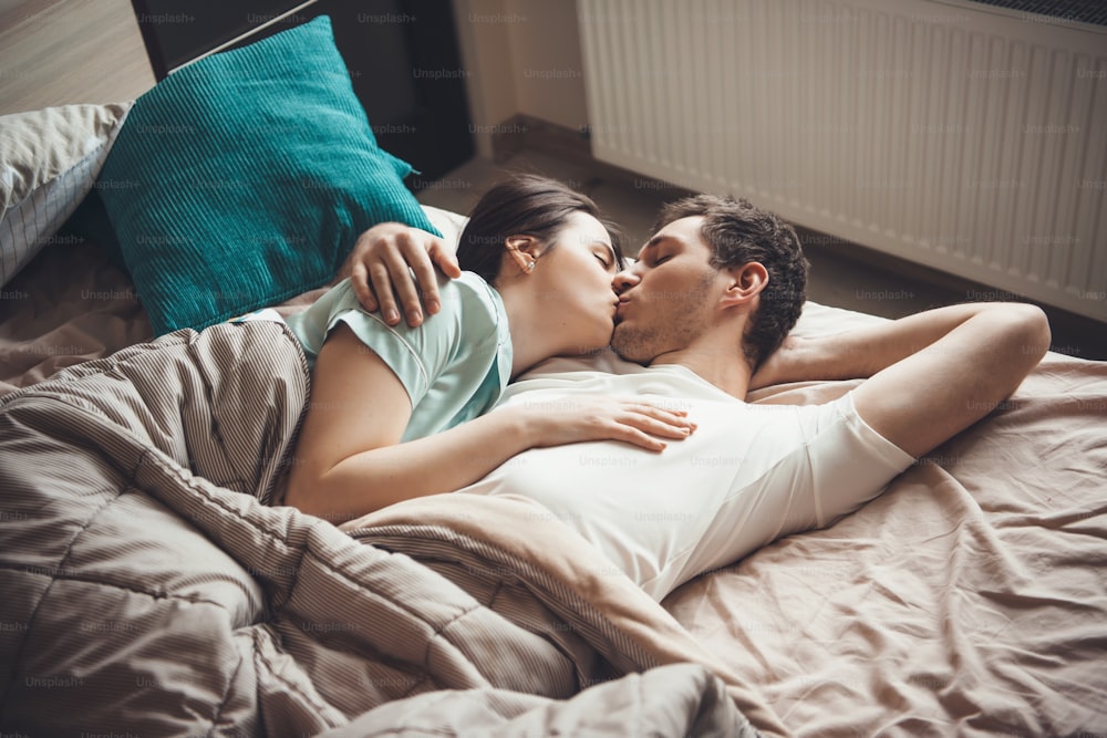 Upper view photo of a young couple lying in bed and kissing while embracing under a quilt