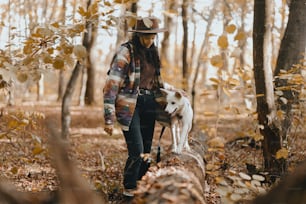 Stylish woman training adorable white dog in sunny autumn woods. Cute swiss shepherd puppy learning with treats. Hipster female with backpack playing with her dog in autumn forest. Space for text