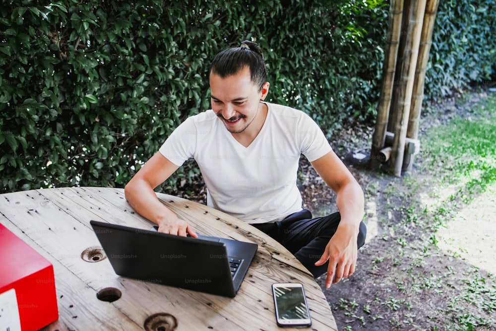 Hombre latino trabajando con computadora portátil al aire libre en una hermosa terraza usando un teléfono inteligente y una computadora sentado con las piernas cruzadas en el trabajo en casa en la Ciudad de México