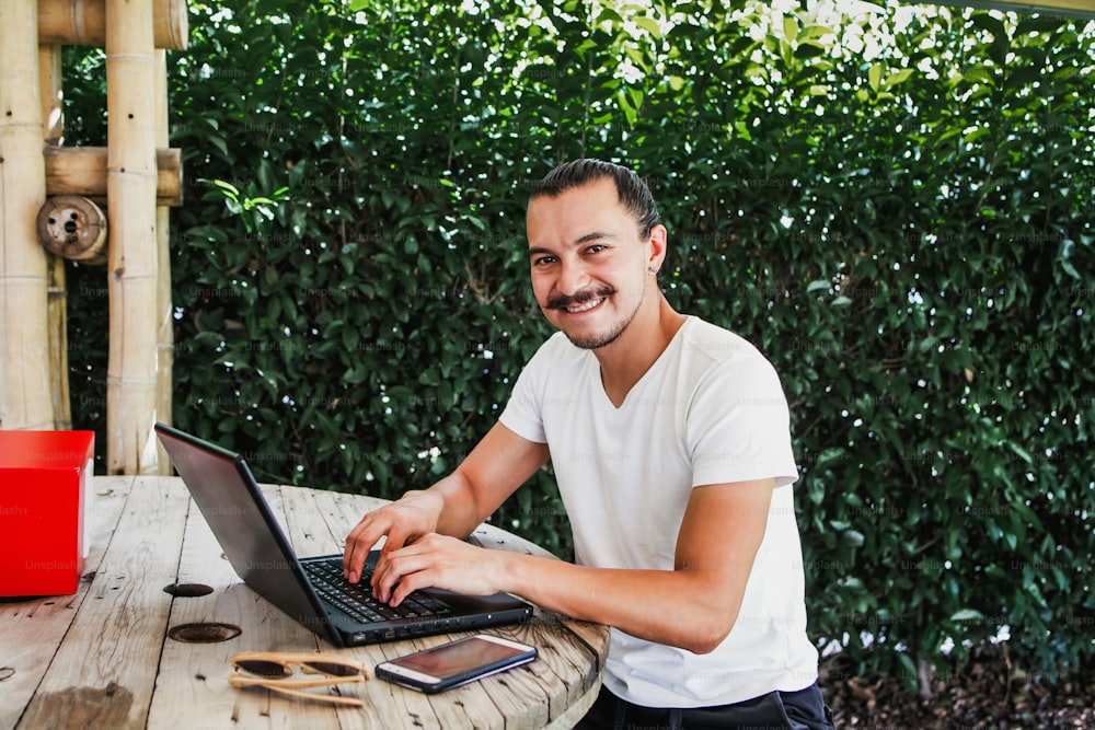 Homme latin travaillant avec un ordinateur portable à l’extérieur dans une belle terrasse à l’aide d’un smartphone et d’un ordinateur assis en tailleur dans le travail à domicile à Mexico
