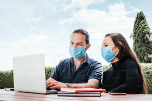 young mexican couple working on laptop with facemask sitting in the rest area of the office outdoors in Mexico for pandemic coronavirus