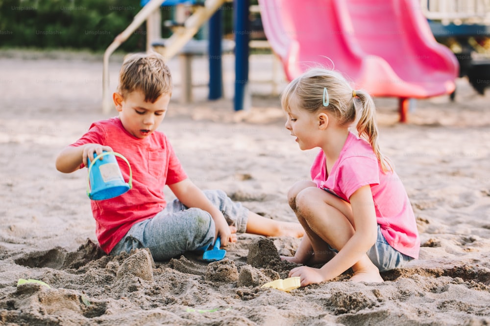 Two Caucasian children sitting in sandbox playing with beach toys. Little girl and boy friends having fun together on playground. Summer outdoor activity for kids. Leisure time lifestyle childhood.