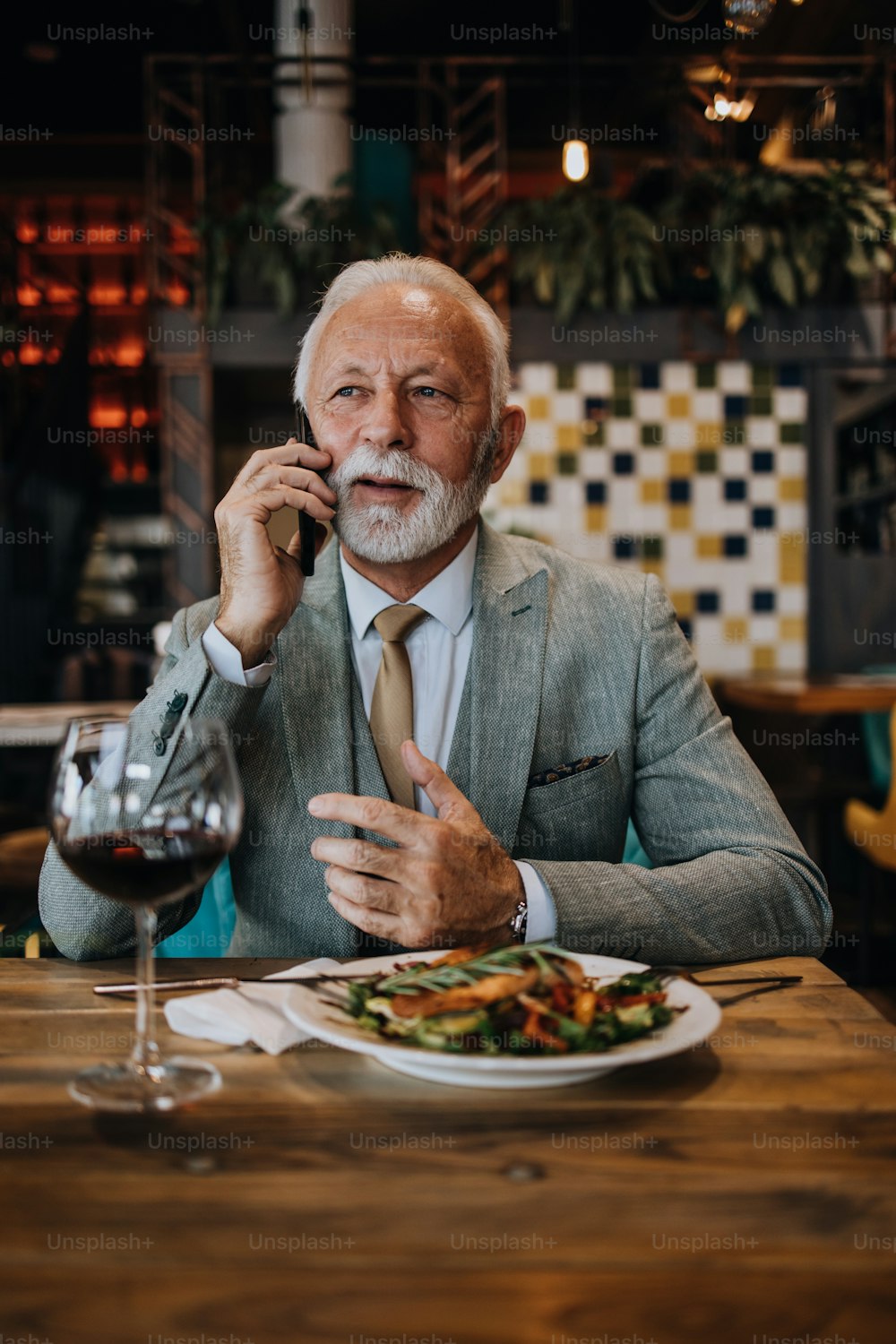Uomo d'affari anziano felice e bello seduto nel ristorante e in attesa del pranzo. Sta usando lo smartphone e sta parlando con qualcuno. Concetto di stile di vita per anziani d'affari.