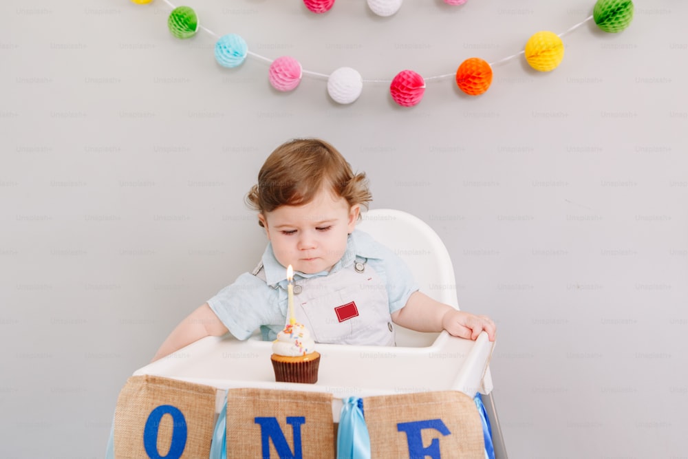 Cute adorable Caucasian baby boy celebrating his first birthday at home. Child kid toddler sitting in high chair eating tasty cupcake dessert with one candle. Happy birthday concept.