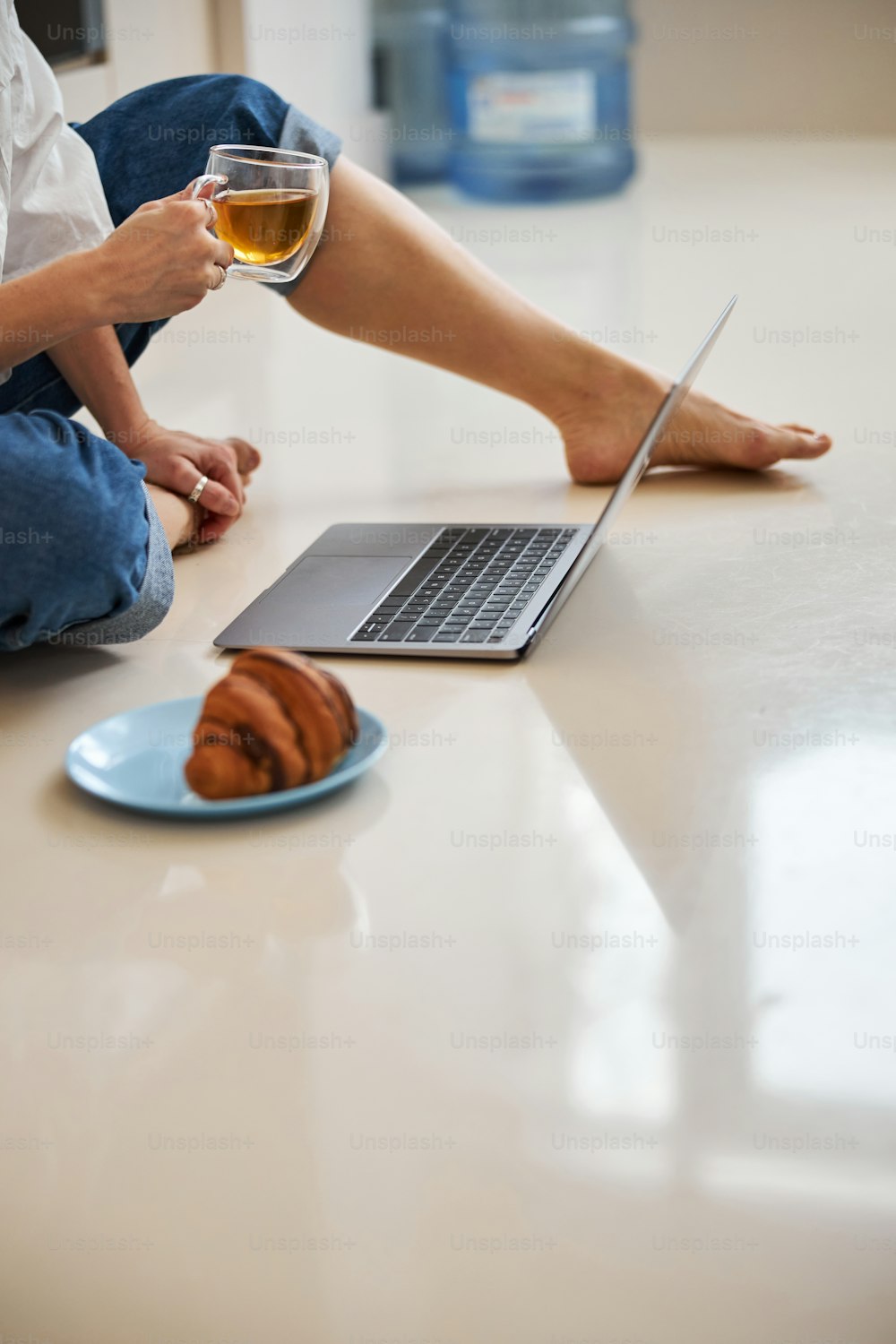 Primer plano de la señora descalza con una taza de té en la mano sentada en el suelo con una computadora portátil moderna y un croissant