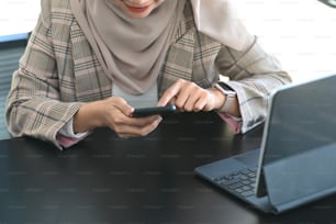 Plan recadré d’une femme musulmane heureuse utilisant un téléphone portable alors qu’elle est assise sur son lieu de travail.
