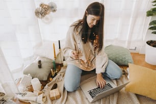 Happy young woman shopping online using laptop and credit card, sitting in stylish boho room with festive holiday decorations. Christmas sale and Black Friday sale and discounts