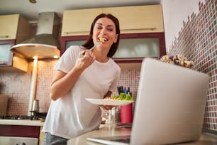 Femmina che porta una fetta di frittata alla bocca aperta mentre trascorre del tempo in Internet con il laptop