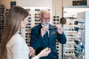 Elegant senior businessman choosing and buying sunglasses in optical store and young female seller helping him to make right decision.