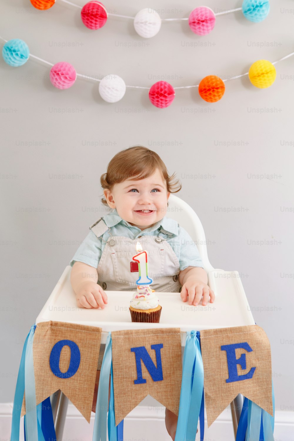 Happy smiling cute Caucasian baby boy celebrating his first birthday at home. Child kid toddler sitting in high chair eating tasty cupcake dessert with lit candle. Happy birthday concept.