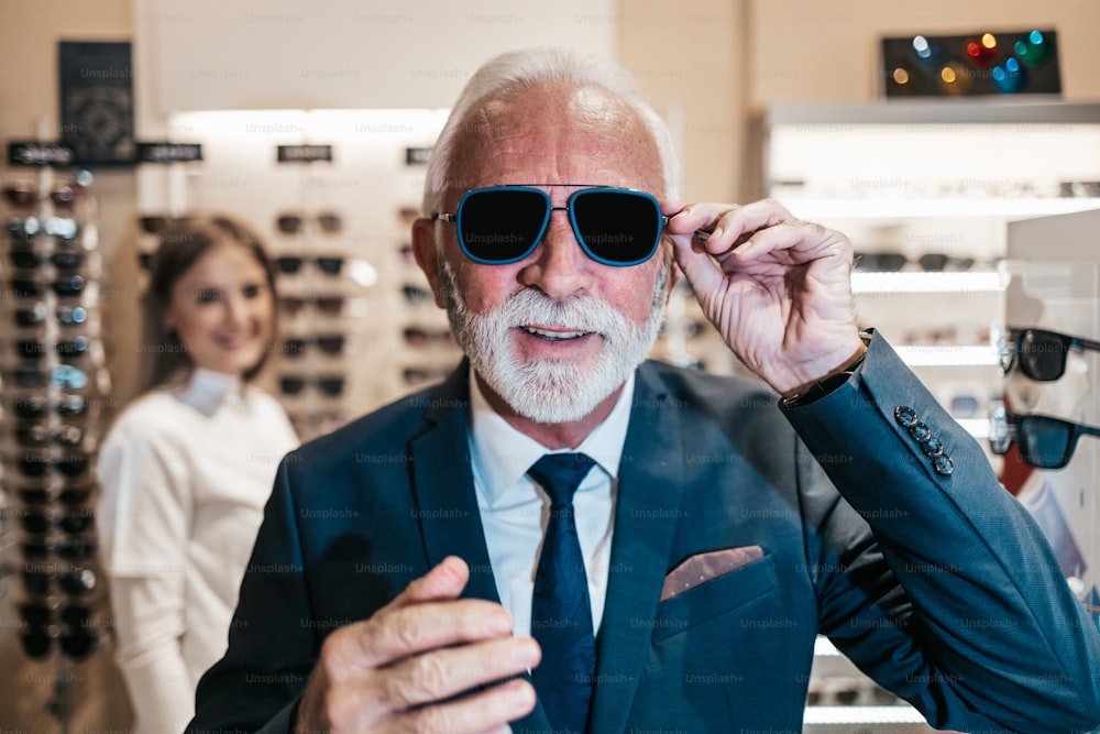 Elegant senior businessman choosing and buying sunglasses in optical store and young female seller helping him to make right decision.