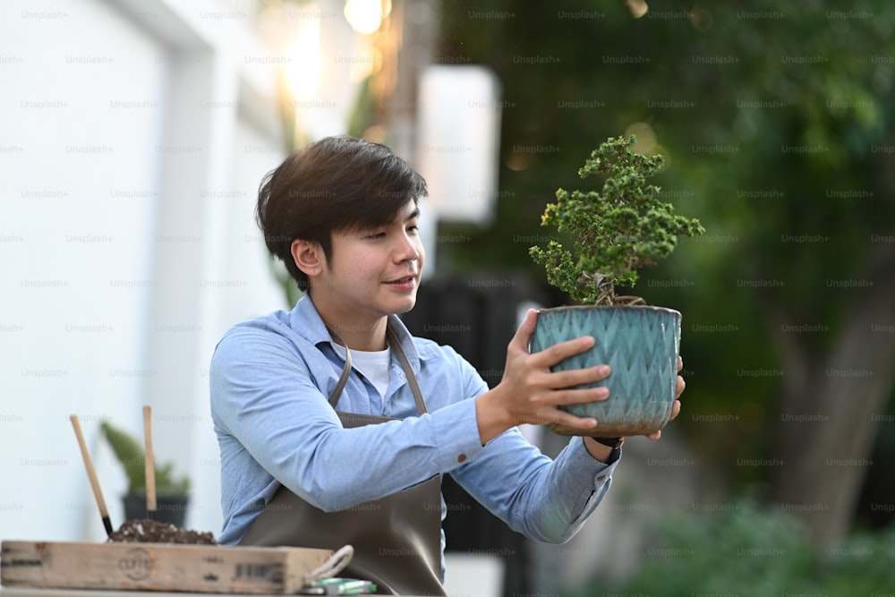 Ein junger asiatischer Mann hält einen Bonsai-Baumtopf in der Hand, während er in seinem Hausgarten sitzt.