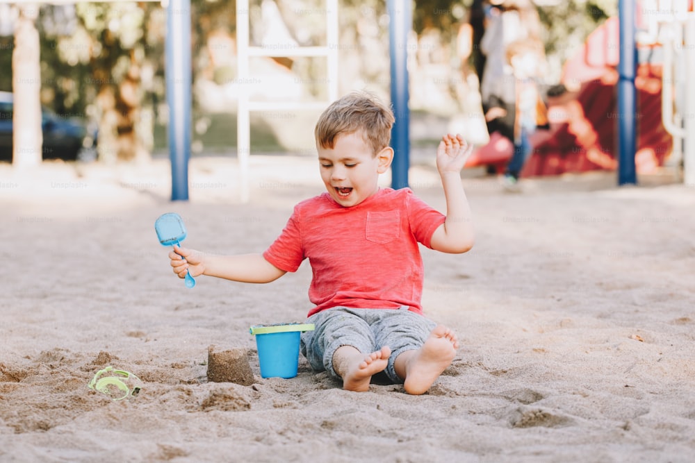 Kaukasisches Kind sitzt im Sandkasten und spielt mit Strandspielzeug. Baby baut Sandburg Sandkuchen. Kleiner Junge hat Spaß auf dem Spielplatz. Sommer-Outdoor-Aktivität für Kinder. Freizeit Lifestyle Kindheit.