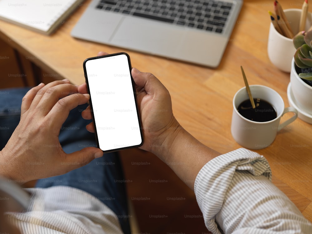 Top view of male hands using smartphone on wooden worktable with laptop, coffee cup and supplies