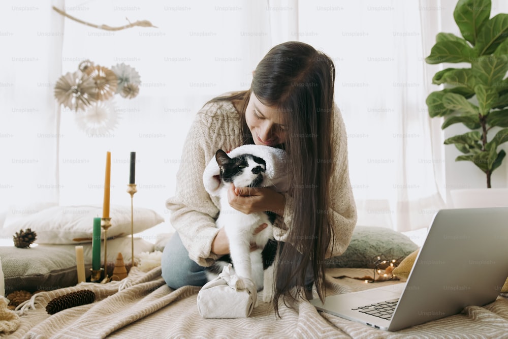 Stylish woman in cozy sweater caressing and kissing cute cat in  santa hat at christmas furoshiki gift and laptop in festive decorated boho room. Celebrating at home, Merry Christmas!