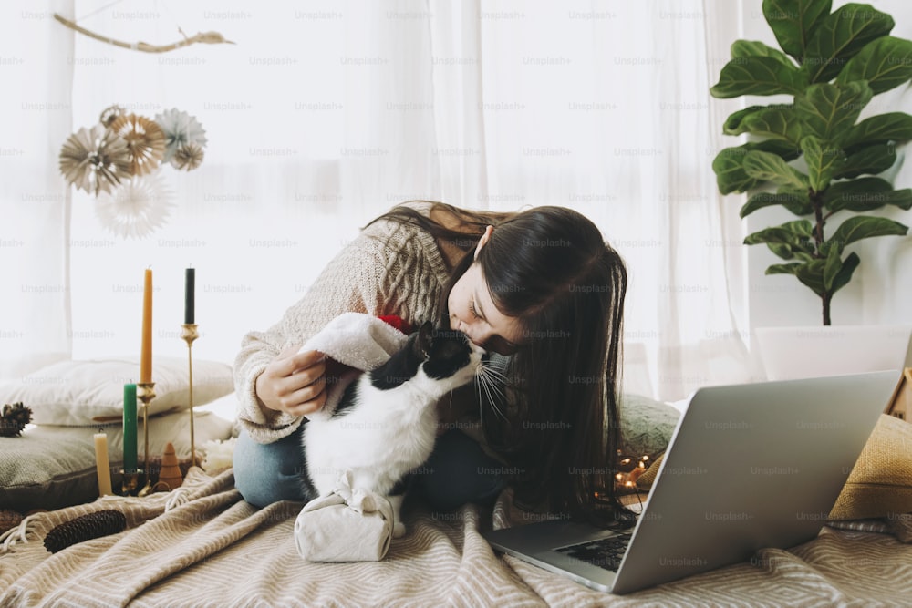 Femme élégante en pull douillet caressant et embrassant un chat mignon dans un chapeau de père Noël lors d’un cadeau furoshiki de Noël et d’un ordinateur portable dans une chambre bohème décorée de manière festive. Célébrer à la maison, Joyeux Noël !