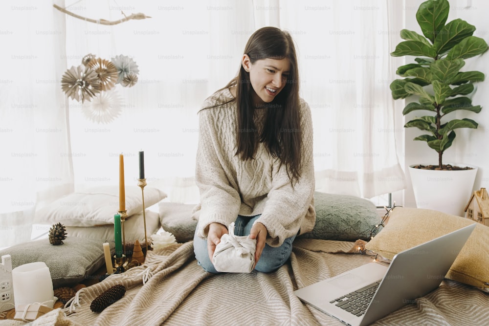 Stylish woman in cozy sweater holding furoshiki wrapped gift in festive decorated boho room and looking at laptop. Young female looking at furoshiki gift wrapping tutorial or chatting online
