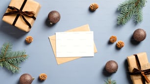 パステルブルーの背景に白紙の紙カードのモックアップ、封筒、クラフト紙のギフトボックス、松の木の枝、クリスマスの飾り。クリスマスや新年のグリーティングカードのコンセプト