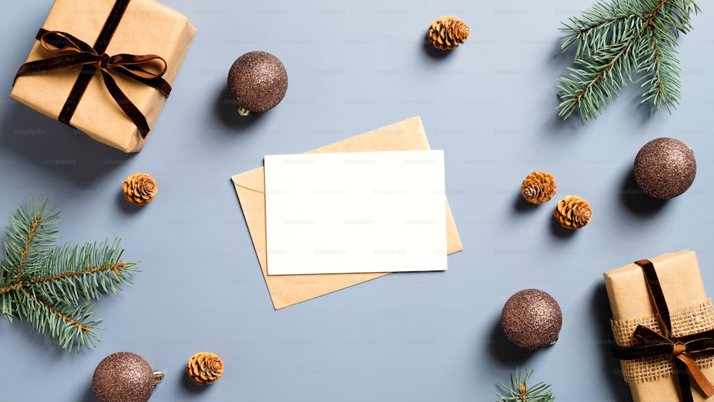 Mockup di carta di carta bianca, busta, scatole regalo di carta artigianale, rami di pino e decorazioni natalizie su sfondo blu pastello. Concetto di biglietto d'auguri di Natale o Capodanno