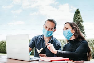 young mexican couple working on laptop with facemask sitting in the rest area of the office outdoors in Mexico for pandemic coronavirus