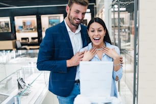 Beautiful couple enjoying in shopping at modern jewelry store. Young woman try it out gorgeous necklace and earrings.