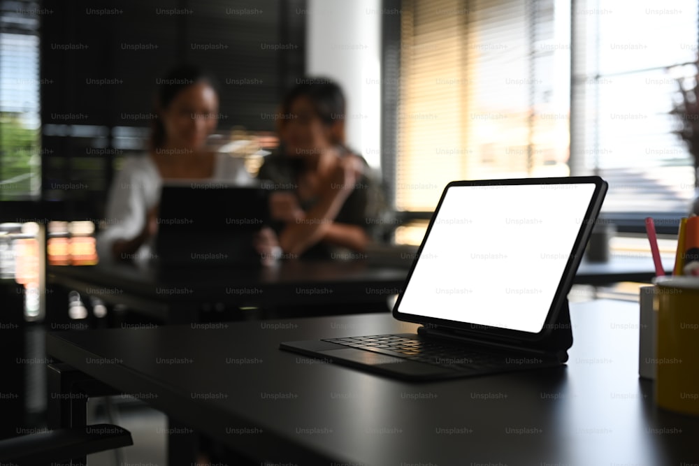 A empty screen computer tablet on black table with two businesswoman sitting in office blurred background.